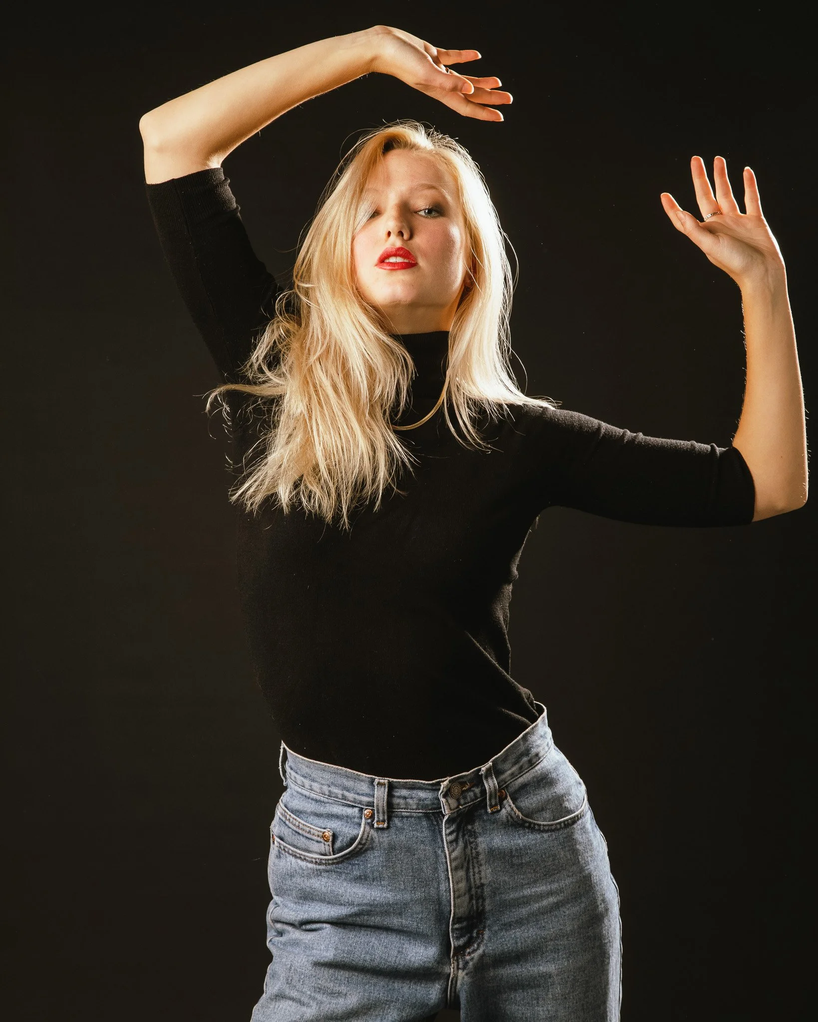 A woman with long blonde hair wearing a black turtleneck and jeans, posing with her arms raised against a dark background.