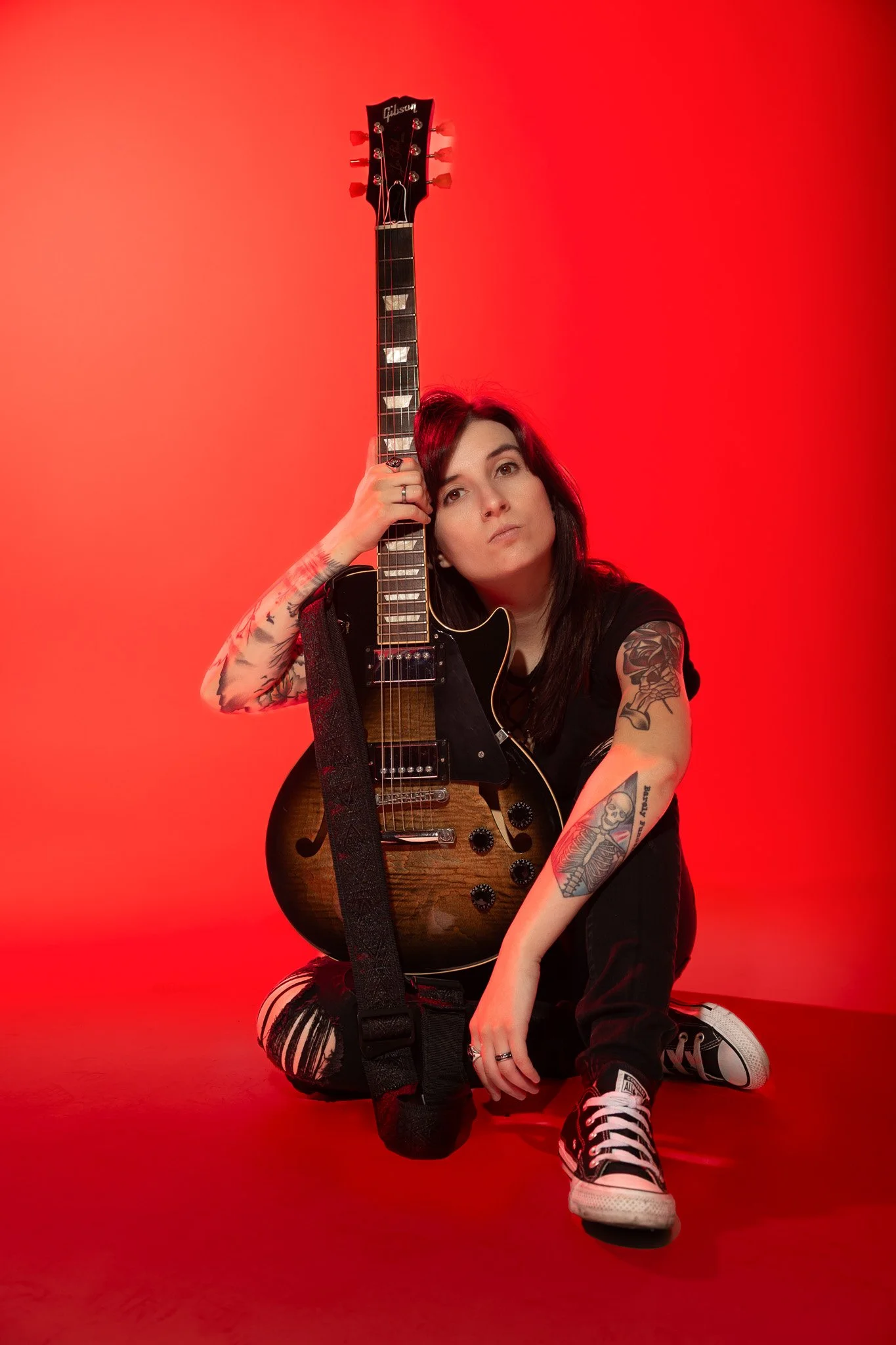 A woman with tattoos sitting on the floor, holding an electric guitar against a red background.