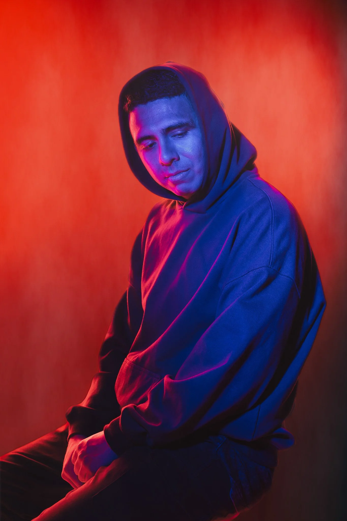 A young man with short dark hair, wearing a dark hoodie, looking down with a contemplative expression, illuminated by pink and blue neon lighting against an orange background.