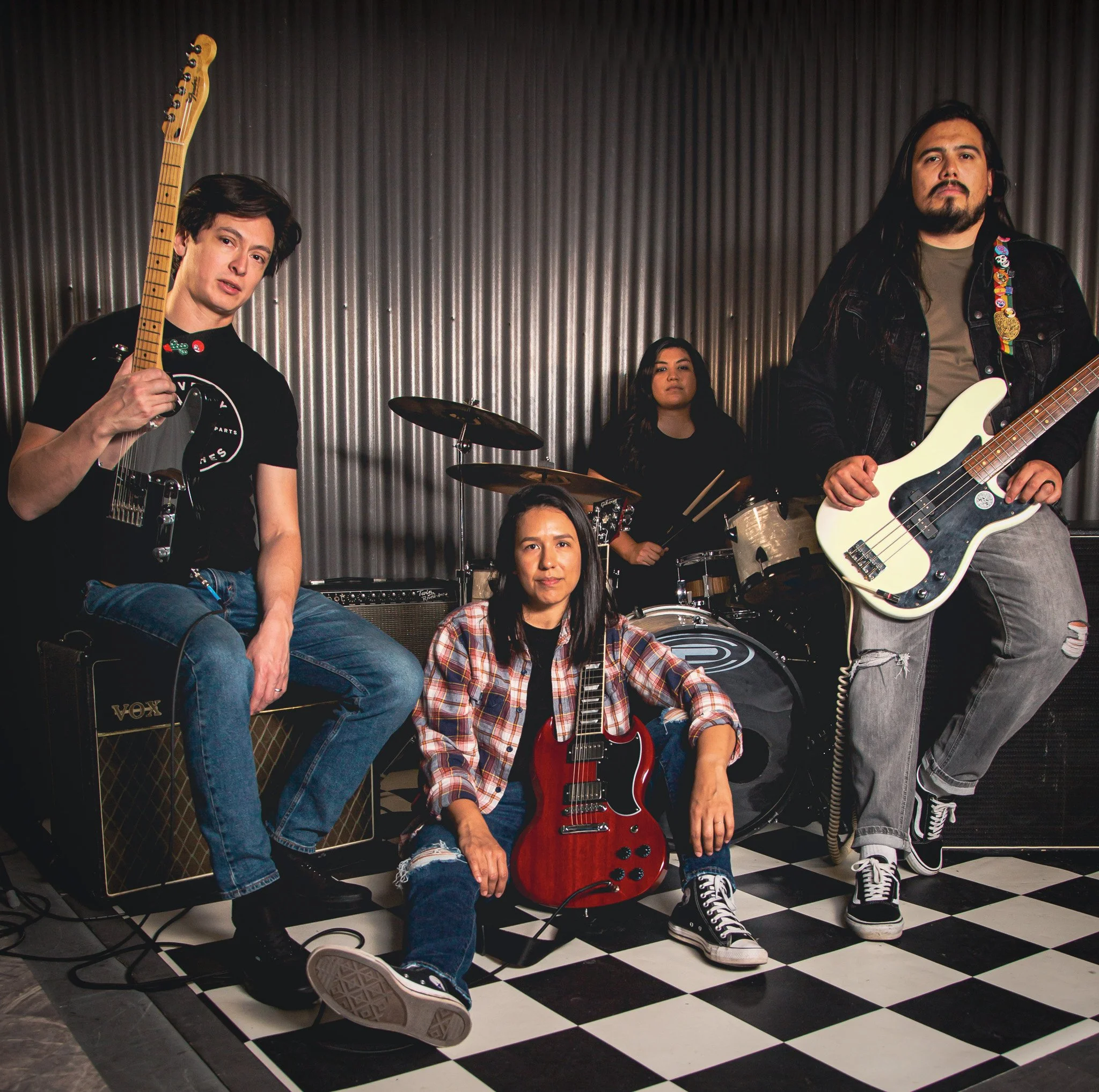 A group of four young musicians with instruments posing in front of a ribbed metal wall. One holds a guitar, another a bass guitar, one sits on a speaker with a guitar, and the fourth is behind a drum set.
