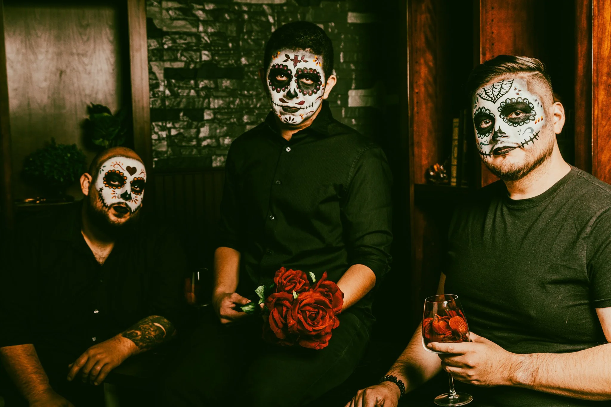 Three men with sugar skull face paint, sitting in a dimly lit room, one holding a bouquet of red roses and another holding a glass of red beverage with rose petals.