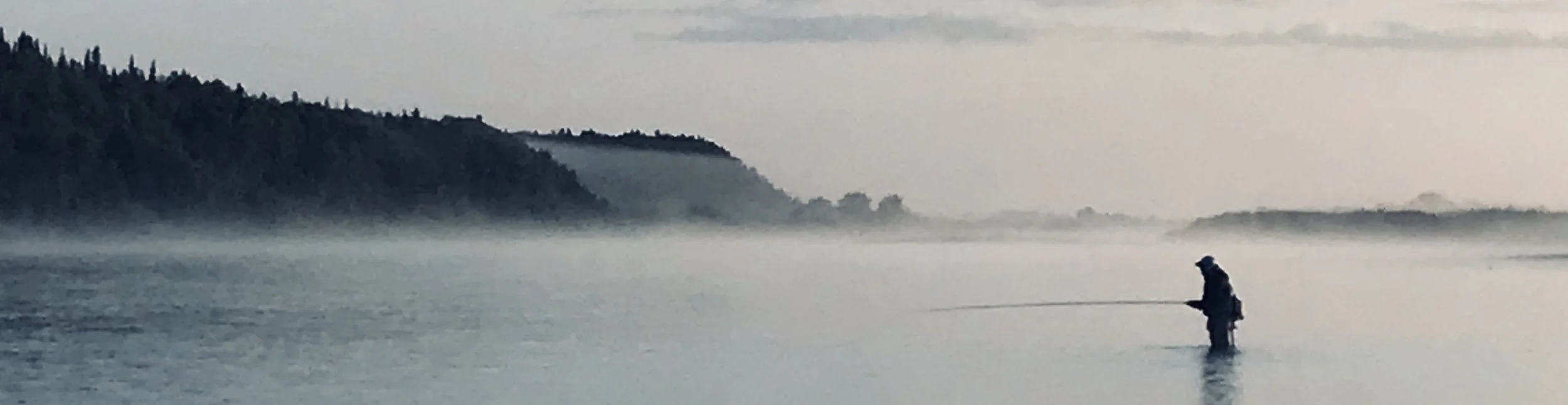 A person fishing in a misty river with a forested hillside in the background.