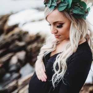 Woman with long blonde hair wearing a green leafy crown, black top, standing outdoors near rocks and water, looking down with a gentle expression.