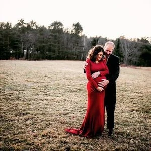 A pregnant woman in a red dress and an older man in a black suit standing together in an open field with trees in the background, smiling and embracing.