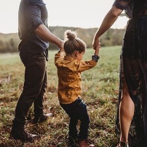 A young girl holding hands with two adults in an open field during sunset, with one adult on each side of her.