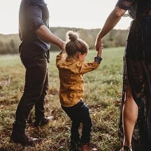 Child holding hands with two adults outdoors in a field.