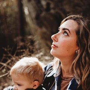 A woman and a young boy outdoors, with trees and sunlight in the background.
