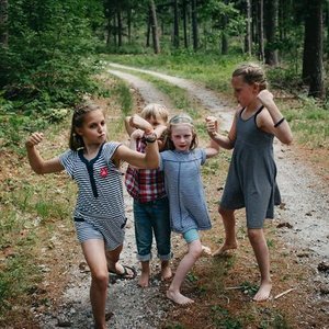Four children and one woman walking on a dirt trail in a wooded area, with some children holding hands and others playing or talking.
