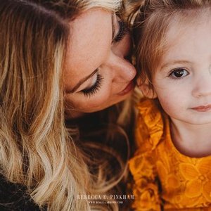 A woman with long blonde hair gently kisses a young girl with blonde hair and a yellow shirt on the cheek, with a blurred background.