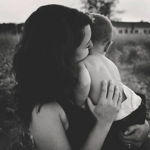 A woman with curly hair is holding a young child close, kissing the child's head in an outdoor setting.