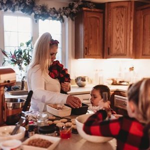 A woman in a white sweater with blonde hair in a kitchen, holding a young girl with a red and black checkered shirt. Two children are at the kitchen counter, one in a red and black plaid shirt, engaged in cooking or baking activities.