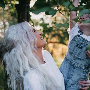 A woman with long, wavy, platinum blonde hair smiling and looking up at a young child who is reaching for a branch in a tree, outside in a park or garden with green leaves and sunlight filtering through.
