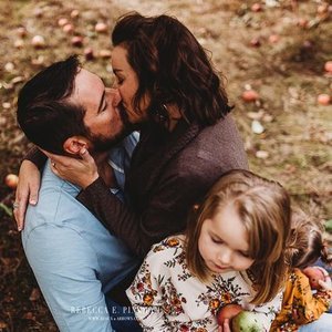 A couple kissing outdoors with a young girl holding a flower in front of them, on a grassy area.