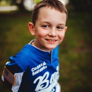 Smiling young boy with short brown hair, wearing a blue sports jersey, outdoors on a grassy field.