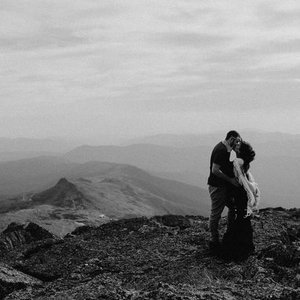 Couple standing on a mountain summit, embracing with a scenic landscape and mountains in the background.