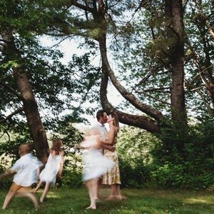 A woman and a girl are kissing in a wooded park, while two other children sit on a swing nearby.