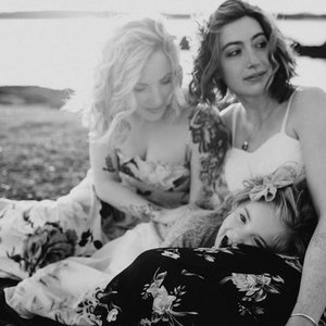 Three women outdoors, one lying down with head on another's lap, all smiling and relaxed.