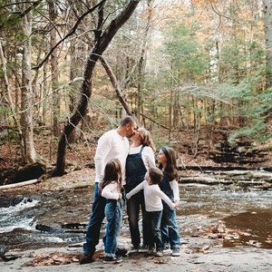 A family of four standing in a creek in a wooded area during fall, with trees in autumn colors surrounding them.