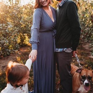 A woman in a long blue dress standing outdoors with a man in a dark jacket and a child with a dog in a natural setting with trees and sunlight.