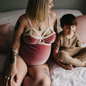 A pregnant woman with tattoos and shoulder-length hair sitting on a bed next to a young boy with dark hair and a tan shirt, both facing sideways.