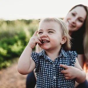 A young girl smiling and touching her face outdoors with a woman, likely her mother, smiling in the background.