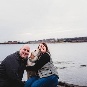 Two people smiling and hugging a dog near a body of water with an overcast sky.