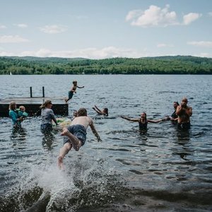 Children and adults swimming and playing in a lake with hills and a partly cloudy sky in the background.