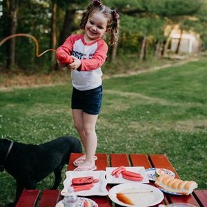 Young girl smiling and playing with a fishing pole on a picnic table outdoors, with a black dog next to her, and food items including bread and slices of meat on the table.