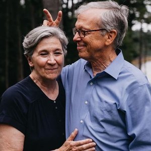 An elderly couple smiling outdoors, with the man making bunny ears behind the woman's head.
