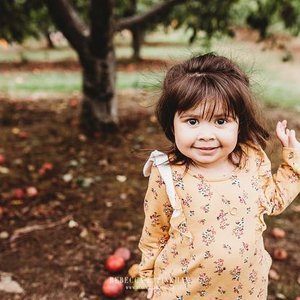 Young girl with brown hair and a floral dress standing outdoors under a tree, smiling, with apples on the ground nearby.