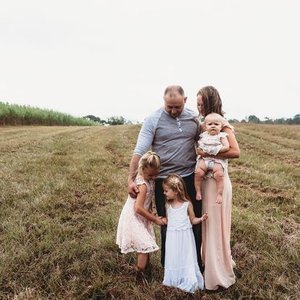A man and a woman standing outdoors with three young girls. The man is holding a toddler, and the women and girls are standing on a grassy field.