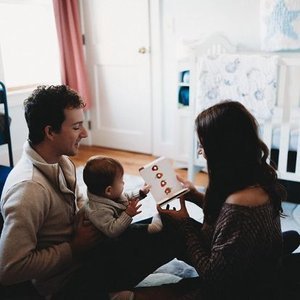 Family of three sitting on the floor in a nursery, looking at a book together.