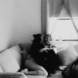 A person sitting on a sofa with a large black dog on their lap, in a room with a window and curtains, black and white photo.