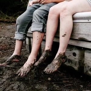 Two people sitting on a dock with their legs dipped in muddy water, with mud on their feet and legs.