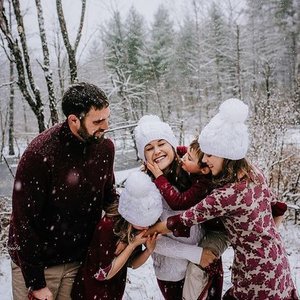 A family of four enjoying a snowy day outdoors, embracing and smiling at each other in a winter forest setting.