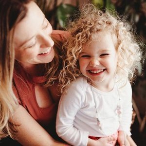 A woman and a young girl with curly blonde hair smiling and laughing together outdoors.