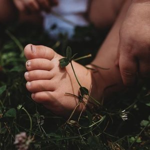 Close-up of a small child's foot with a green insect sitting on toes, surrounded by grass and natural outdoor setting.
