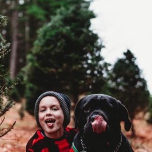 A young boy with a beanie smiling and sticking out his tongue, sitting next to a black dog with its tongue out, outdoors in a wooded area.