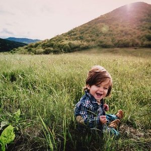 Young boy with brown hair sitting on the grass in a field, smiling and laughing, with green hills and mountains in the background.