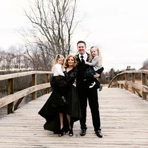 A family of four standing on a wooden bridge outdoors, with trees and cloudy sky in the background. The adults and children are dressed in formal clothing.