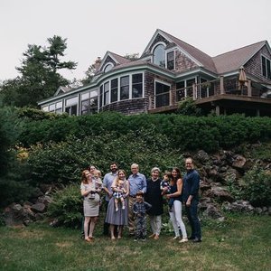 Group of eight people standing on grass in front of a large house with a balcony and trees.