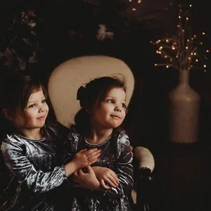 Two young girls in matching velvety gray dresses sitting closely together in a dimly lit room, with warm lighting and decorative vases in the background.