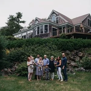 Group of seven people standing on a grassy lawn in front of a large house with a wraparound porch and multiple stories, surrounded by trees and rocks.