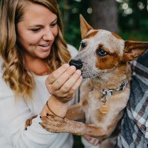 Woman smiling while holding a small dog’s paw, outdoors in a park.