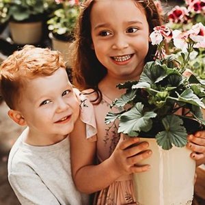 A young girl holding a potted pink flower plant, standing next to a young boy with curly red hair in a greenhouse or garden center.