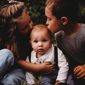 A woman and a young boy showing affection to a baby, who is sitting on the grass. The woman kisses the baby on the cheek, and the boy leans in close to the baby's face.