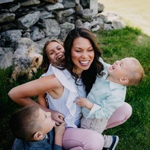A woman with two young children and a girl, laughing outdoors on green grass near rocks, enjoying a joyful moment.