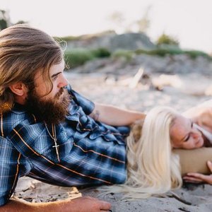 A man with a beard lying on the sand next to a woman with blonde hair, on what appears to be a beach.