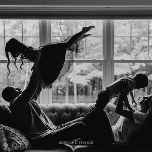Person lying on a couch in a living room, lifting a child into the air against a large window background.