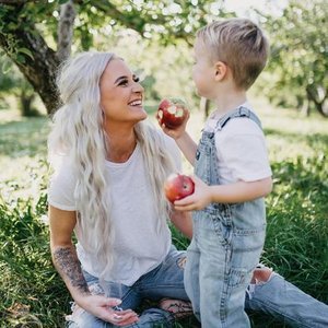 A woman with long, light-colored hair sitting on the grass outdoors, smiling at a young boy holding two red apples, with trees and greenery in the background.